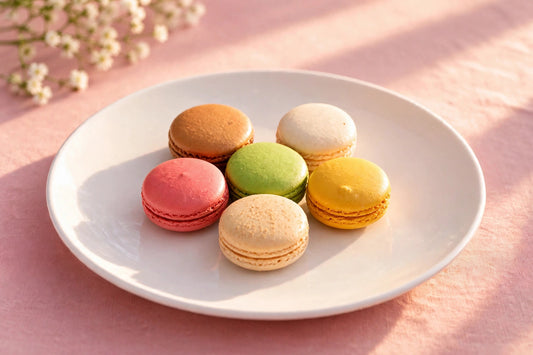Six colorful La Crème Bakery macarons on a white plate over a soft pink linen background
